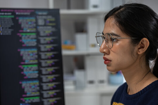 Young Asian woman wearing eyeglasses viewing computer code on monitor with focused expression in modern office workspace