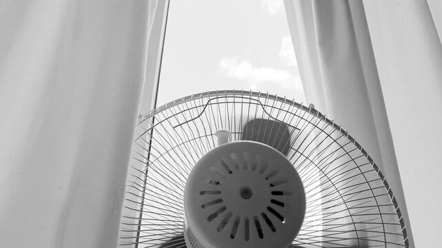 Monochrome footage of an electric floor fan blowing air indoors by a bright window with sheer curtains