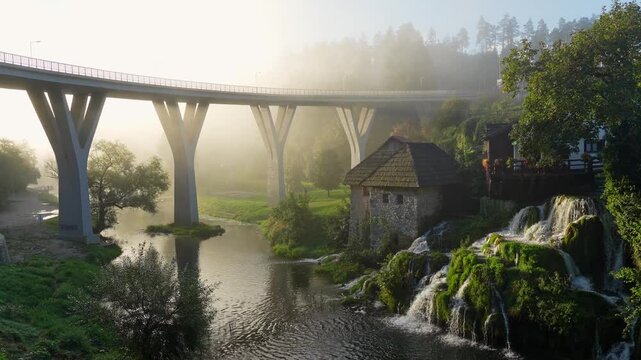 Slunj Rastoke aerial view featuring historic stone house by waterfall and modern bridge at sunrise.