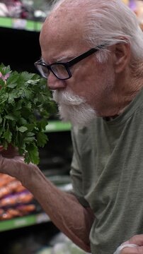 An elderly man shops in a grocery store, carefully choosing fresh herbs in the produce section.
