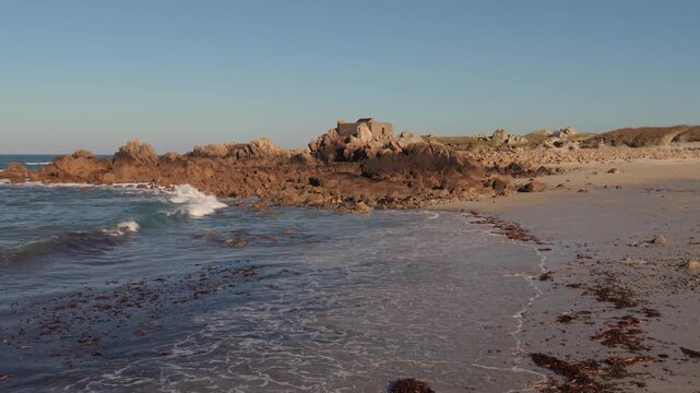 Scenic coastal landscape with tranquil ocean waves rolling onto sandy shore and ancient stone ruins located on rocky headland.