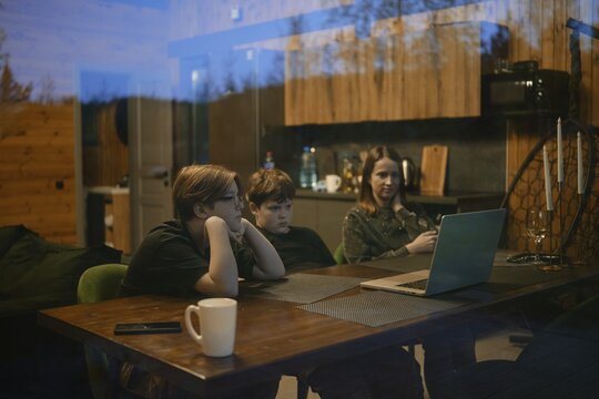 Inside a cozy cabin, a family gathers around a table enjoying a shared experience. Two children are focused on a laptop, while an adult casually checks a phone, creating a warm atmosphere