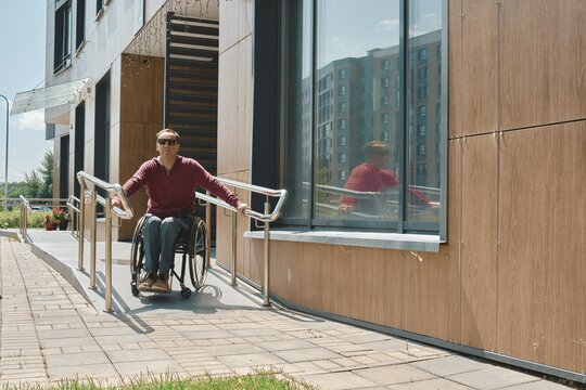 Individual in a wheelchair is utilizing a ramp to exit a modern building with large windows. The bright sun illuminates the exterior, creating a vibrant urban atmosphere