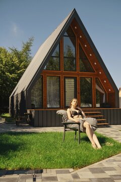 Carefree woman in bikini sunbathing on lawn at contemporary A-frame house with large glass windows and a triangular facade