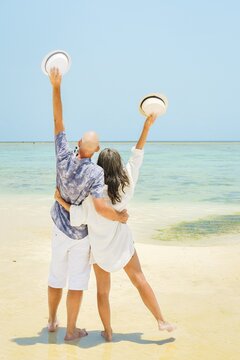 Couple stand together hugging hug enjoy arms up on white sand beach in sunny tropical destination. Romantic summer holiday by turquoise ocean with clear blue sky. Honeymoon paradise vibes