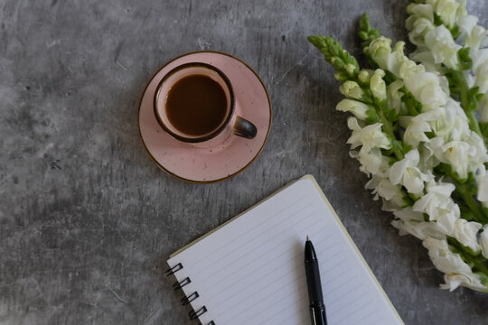 Pink saucer holds dark coffee. White flowers curve beside a notebook. Black pen rests open pages. Concrete background adds textured calm.