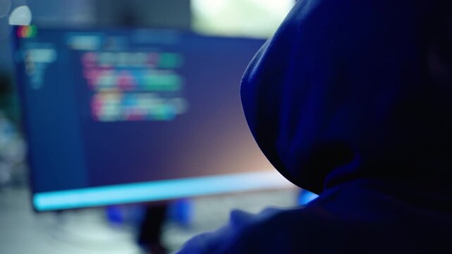 Anonymous hacker in a dark hoodie typing code on a computer screen. Cybersecurity threat, cybercrime, and software development concept. Blue neon lights illuminate the dark room at night.