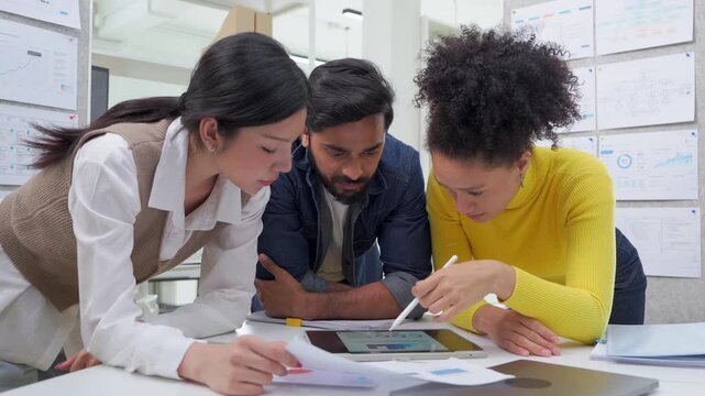 Three colleagues lean over a desk covered in printed charts and a tablet, closely reviewing data together during a focused analysis session in a modern office lined with project boards.