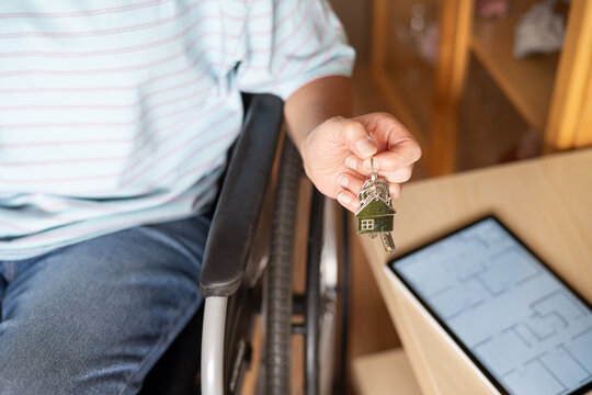 Person in a wheelchair holding keys with a house-shaped keychain, symbolizing home ownership, independence, and the concept of accessible housing while looking at a floor plan on a tablet