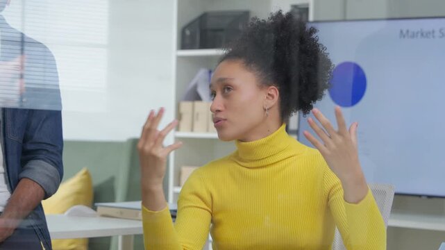 A confident businesswoman in a yellow sweater gestures while speaking, with a colleague reflected through a glass wall in the background of a bright, modern office setting	