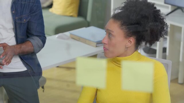 A confident businesswoman in a yellow sweater gestures while speaking, with a colleague reflected through a glass wall in the background of a bright, modern office setting	