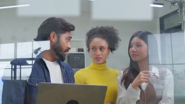 A confident businesswoman in a yellow sweater gestures while speaking, with a colleague reflected through a glass wall in the background of a bright, modern office setting	