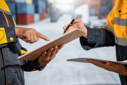 Two warehouse workers in safety uniforms review and sign a clipboard during a shipment inspection in a blurred industrial storage area.