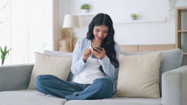Happy young asian woman relax on comfortable couch at home texting messaging on smartphone, smiling girl use cellphone, chatting online message, shopping online from home