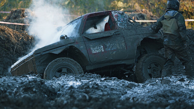 Mud splattered racing car in thick sludge, steam rising from engine, driver in helmet gear standing nearby
