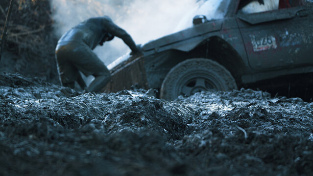 Low angle view of person pushing muddy off-road vehicle stuck in thick mud during racing. Cool blue tones dominate. Shallow depth of field focuses on foreground mud texture