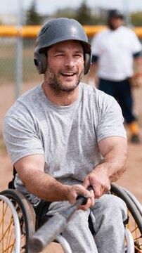 Male athlete in wheelchair baseball uniform smiles while gripping bat, playing on outdoor field with blurred teammates in background during a para sport event