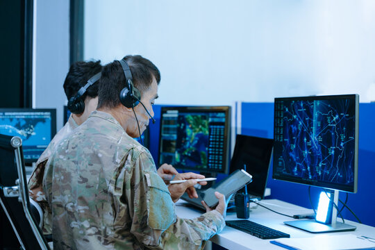 Senior military officer in camouflage using a tablet to analyze tactical radar data alongside his team in a modern cybersecurity command center.