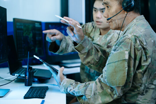 Two military officers in camouflage uniforms analyze tactical cybersecurity data on a tablet and multi-monitor displays