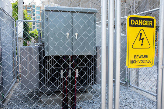 Electrical cabinet stands behind chain link fence, yellow danger sign warns of high voltage hazard nearby, secure enclosure protects power equipment at outdoor industrial installation site