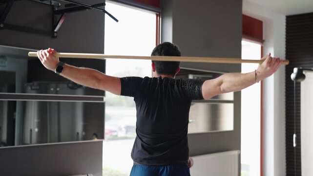 Athletic man practicing shoulder mobility exercises with wooden stick in modern gym.