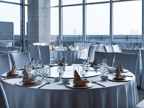 Luxury hotel banquet dining area featuring round tables with white tablecloths, formal place settings, white chair covers, floor-to-ceiling windows, patterned carpet