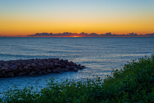 Golden sunrise over a calm blue sea with a rocky breakwater and green coastal vegetation, vivid orange sky glowing above dark clouds on the horizon, a peaceful and inspiring seascape in dawn light