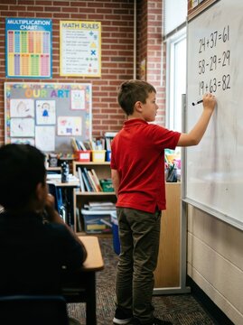 Elementary school student writing addition and subtraction equations on a whiteboard in a bright classroom