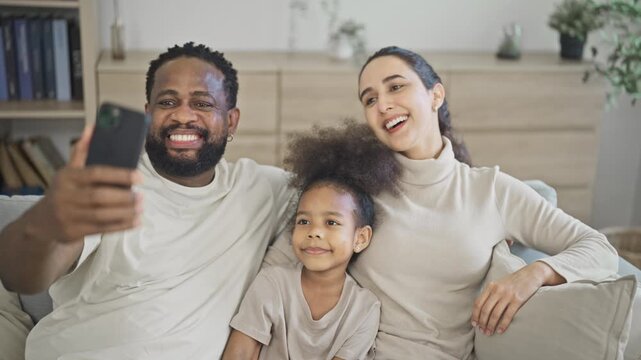 Multiracial family of three smile brightly at the camera as the father holds up a smartphone for a family selfie on the sofa in a warm modern home living room.