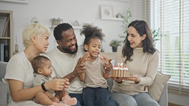 Extended multiracial family including a mother holding a newborn clap and smile as a young girl blows out her birthday candles in a bright modern living room.