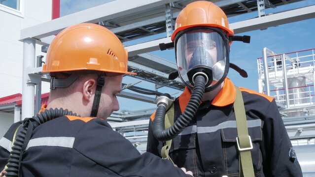 Two male industrial workers in orange helmets wear respirators at plant, one facing camera with focused expression. Safety gear and steel framework suggest industrial style at refinery or factory
