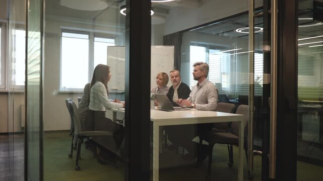 A view through a glass partition shows a modern, busy office interior. Job interview process is in office. Human resources, meeting and group of hiring candidates for vacancy