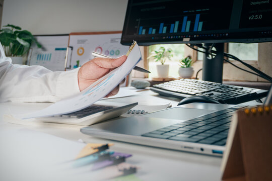 Business analyst reviewing printed charts, financial reports while taking notes at modern office desk, representing data analysis, financial planning, performance evaluation, strategic decision making