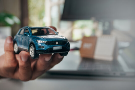 Blue toy car held in hand with blurred home office background, representing vehicle ownership, automotive insurance, car financing, transportation planning, and personal financial decision making