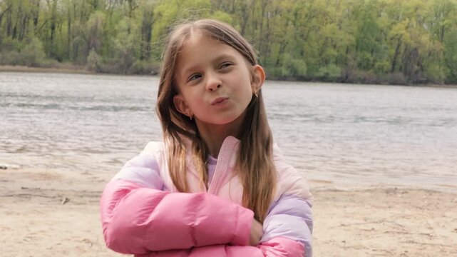 Little girl in puffer jacket playing and gesturing by the river on a spring day