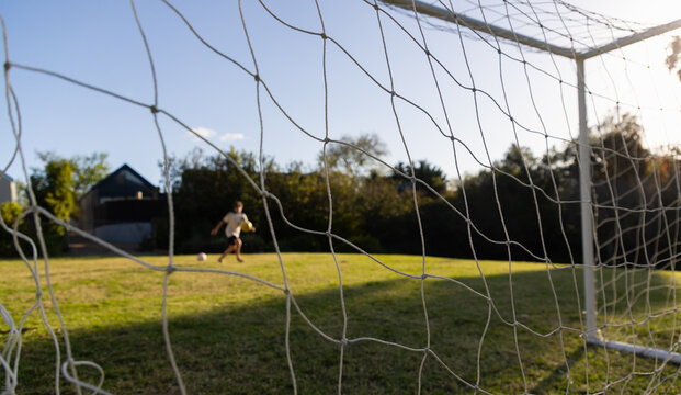 soccer net view of teenage boy striking soccer ball into net at sunset