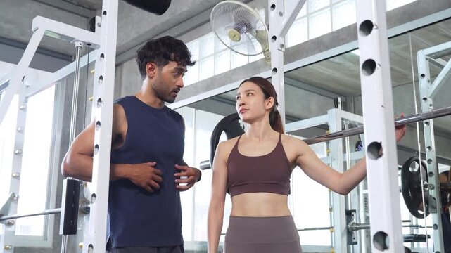 Trainer coaches a woman during a strength training session in a modern gym. Discuss exercise technique beside a squat rack and weight equipment, fitness coaching, motivation, healthy lifestyle.