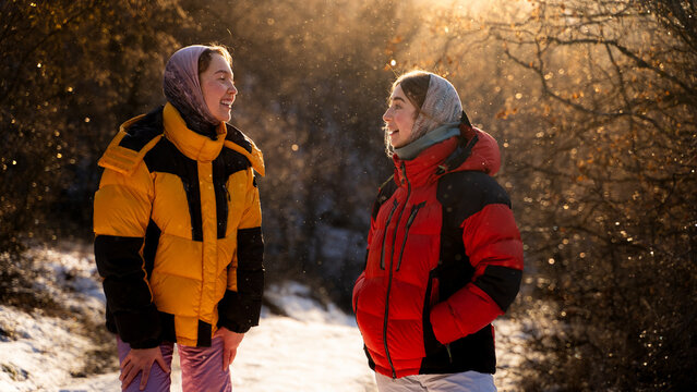 Two women enjoying a friendly conversation during a winter walk in a snowy park surrounded by warm sunlight. Friendship, companionship and outdoor lifestyle.