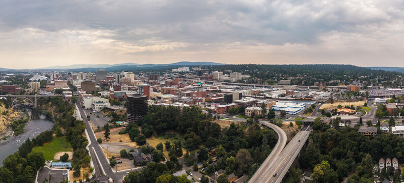 Spokane, Washington, United States - 15 August 2026: Aerial view of the Spokane city skyline with the Spokane River, Monroe Street Bridge, urban buildings, and forested hills under a cloudy sky.