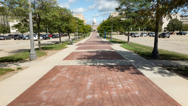 Lansing, Michigan, United States - 25 August 2026: Aerial view of Michigan State Capitol with a brick walkway, trees, and parking lots under a blue sky.
