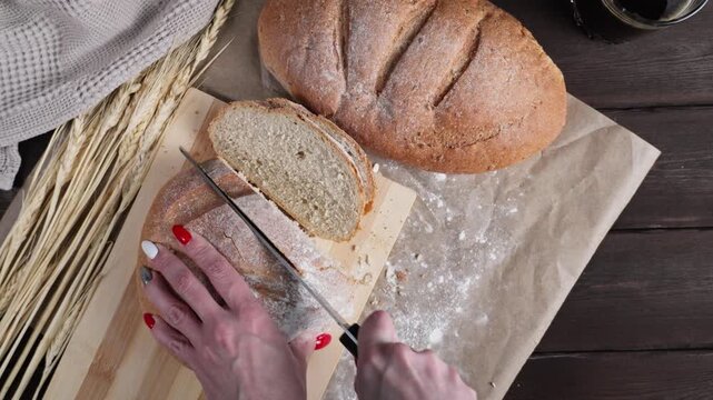 Close-up of a woman slicing fresh rustic bread into pieces with a serrated knife, surrounded by wheat stalks, sliced bread and a dark beverage