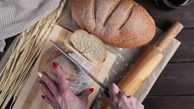 Close-up of a woman slicing fresh rustic bread into pieces with a serrated knife, surrounded by wheat stalks, sliced bread and a dark beverage