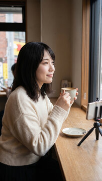 A serene young Japanese woman sitting by a bright wooden cafe window, smiling and holding a ceramic cup of coffee while her smartphone is mounted on a small tripod for a video call.