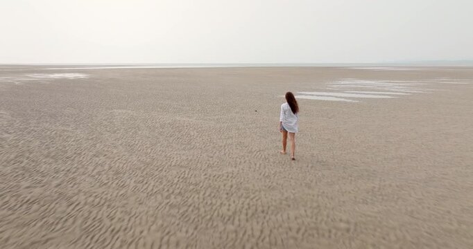 Beautiful woman walking barefoot along vast low-tide beach in Ko Lanta, Thailand, back to camera, serene seascape and open horizon, peaceful solitude, gentle rippled sand textures. Aerial drone flight