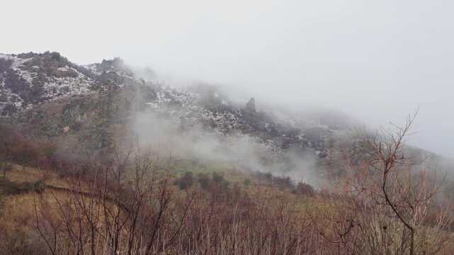 Misty mountain landscape in early spring with patches of snow, bare trees and rocky slopes partially covered by drifting fog