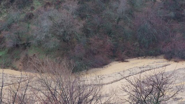 Powerful mudflow rushes down a mountain slope, carrying muddy water and debris over a damaged road after heavy rainfall, illustrating a natural disaster and infrastructure damage