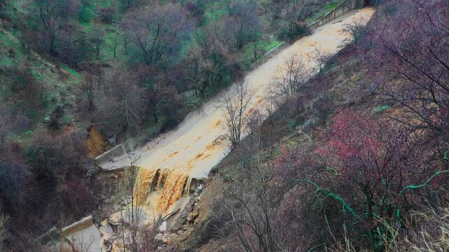 Powerful mudflow rushes down a mountain slope, carrying muddy water and debris over a damaged road after heavy rainfall, illustrating a natural disaster and infrastructure damage
