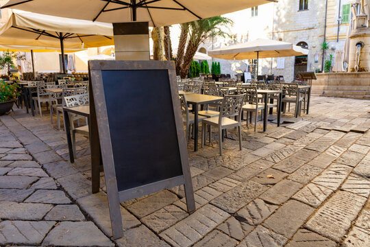 Black chalkboard, empty blank restaurant menu blackboard on street near chairs and tables of a cafe. Black menu chalkboard mockup, menu board template