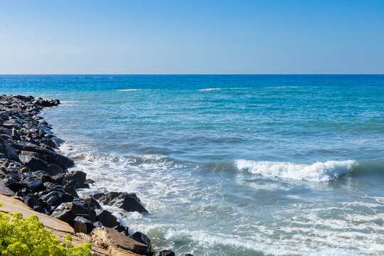 Blues waves crash against the rocks, creating a vivid and energetic coastal scene. The bright sky and deep blue sea blend beautifully