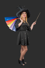 Smiling woman wearing a black witch hat and dress, carrying colorful shopping bags on her shoulder while holding a magic wand, blending holiday spirit with retail therapy © somemeans